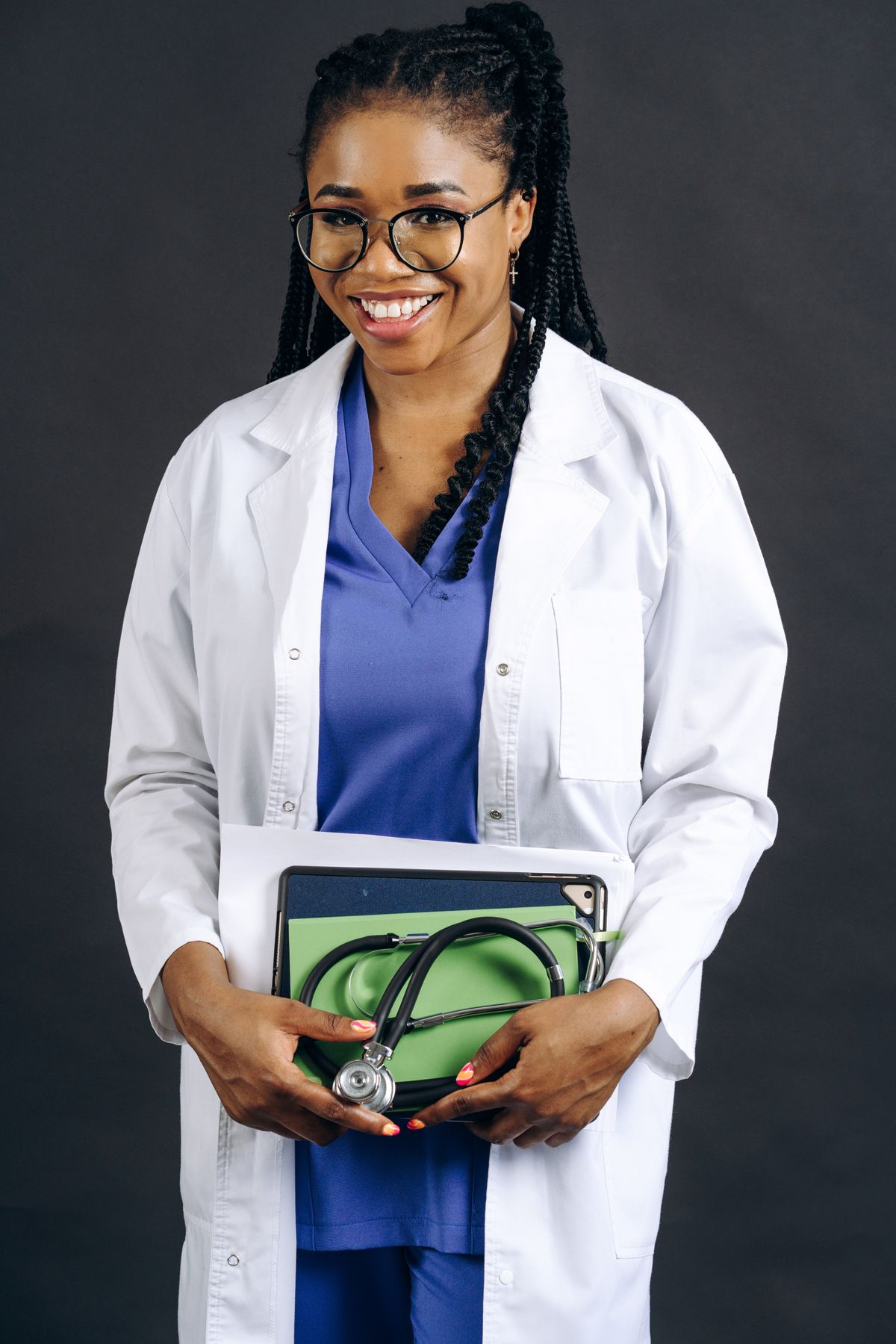 A smiling healthcare professional wearing glasses and a white coat, holding a stethoscope and tablet
